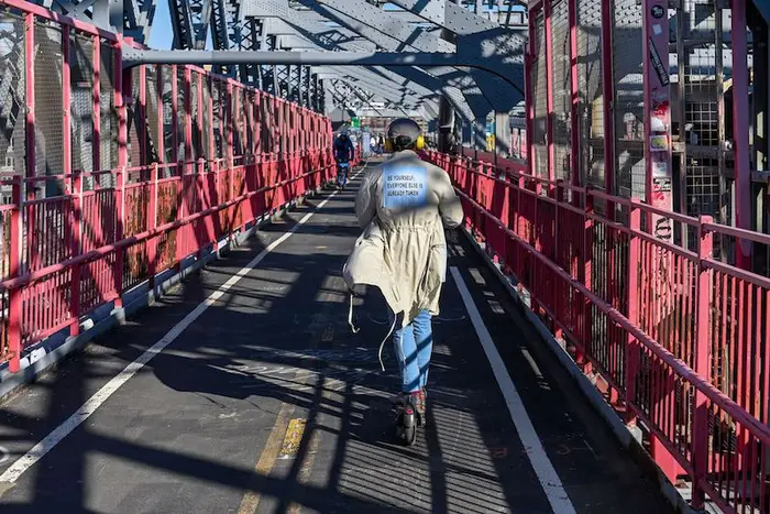A person with back to camera wearing a coat that reads "Be Yourself" rides an e-scooter over the Williamsburg Bridge on a sunny day.